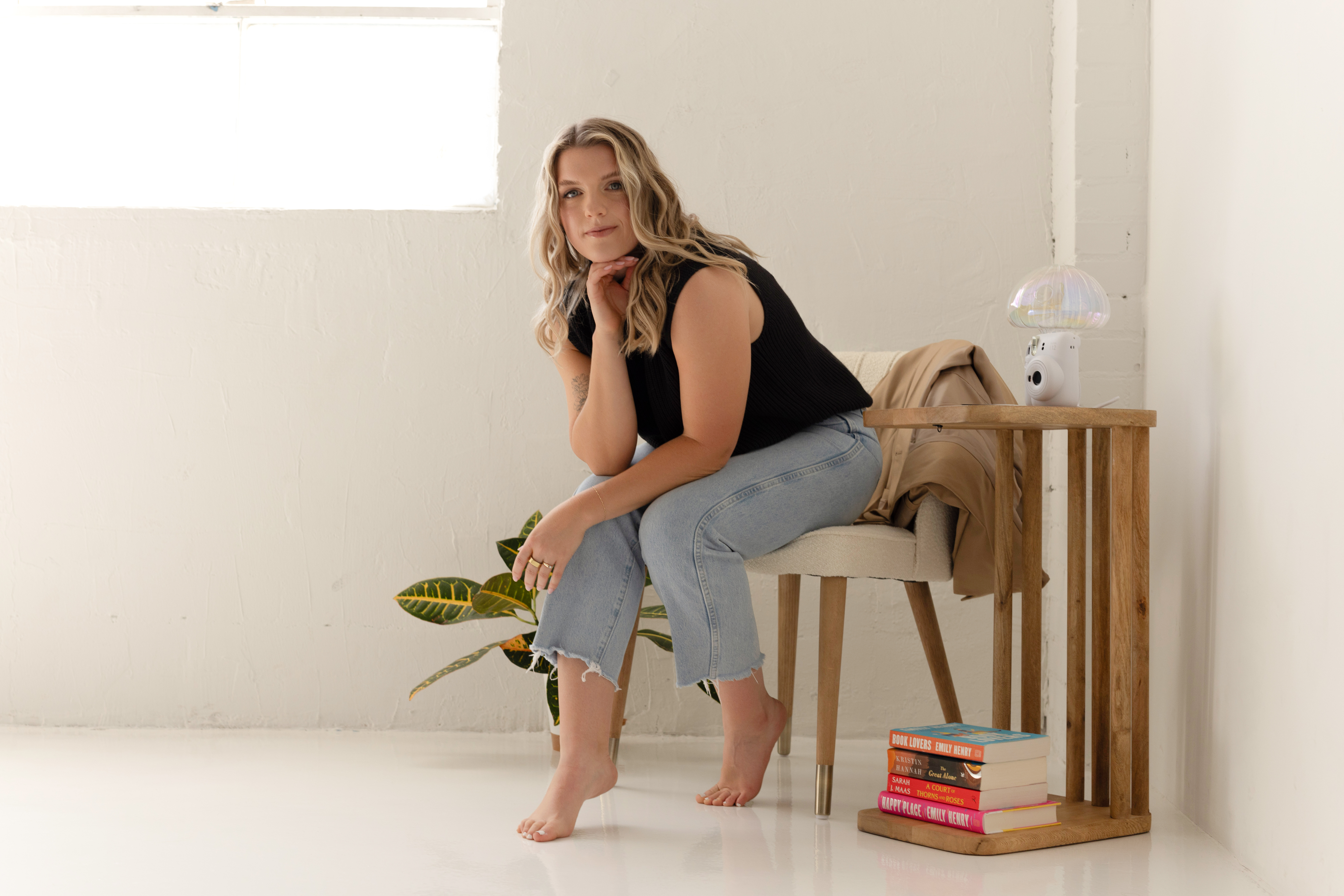 How to Increase Your Inquiries During a Slow Season | Reveal Studio Co Kaili Meyers wearing a black top and light jeans sitting on a armless chair against a white brick background.